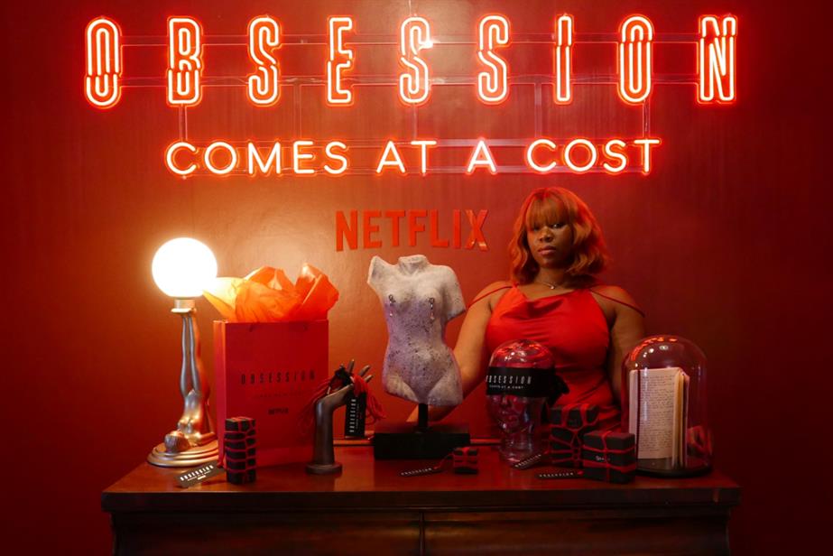 Woman dressed as Anna standing with sex products and neon sign that says Obsession comes at a costt